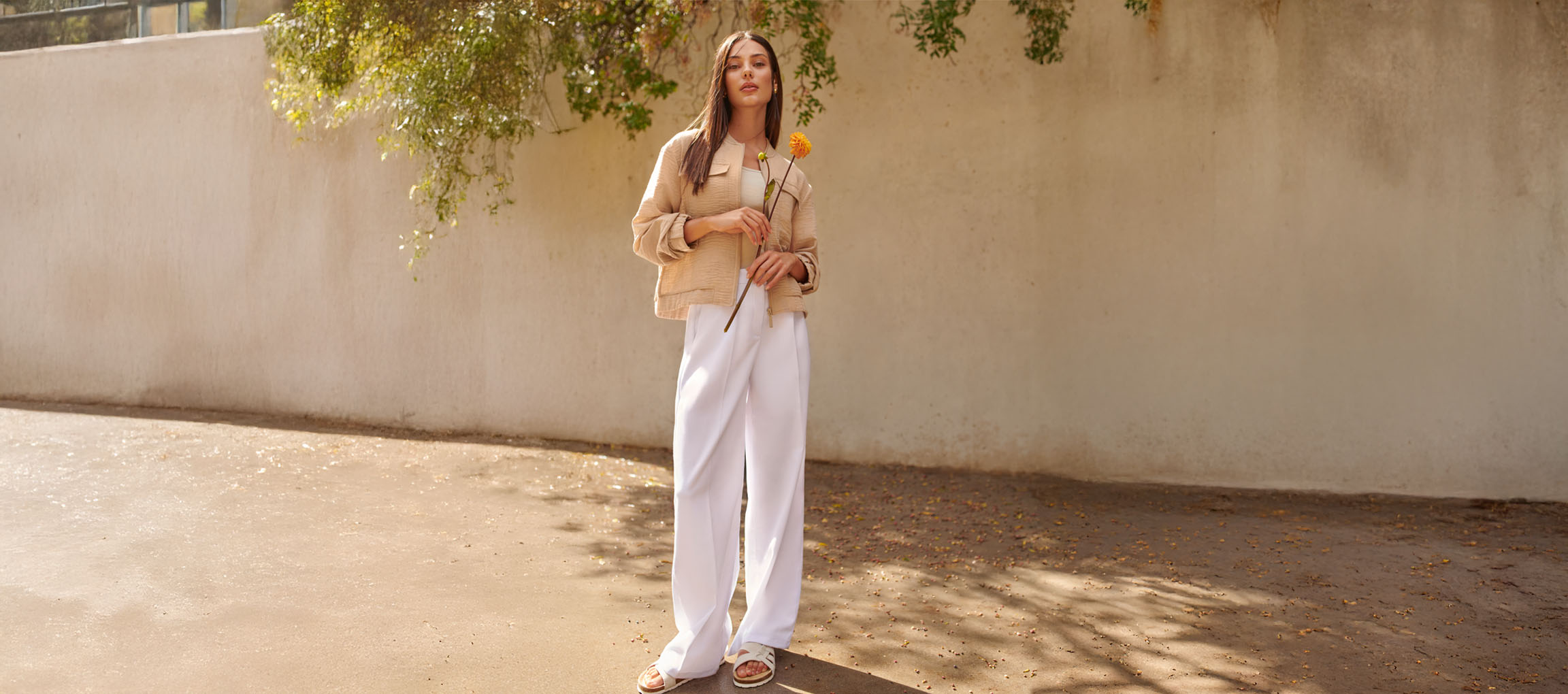 Nainen beigessä takissa, valkoisissa housuissa ja sandaaleissa, pitäen keltaista kukkaa.