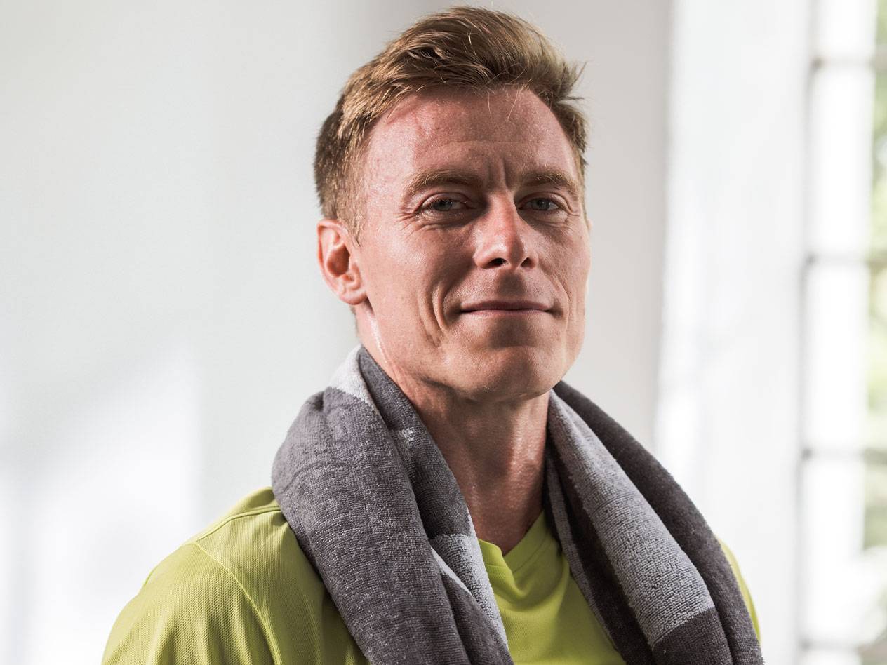 Vaaleahiuksinen mies, jolla on pyyhe kaulassaan, katsoo kameraan.