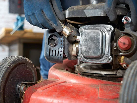 Käsineet kädet vaihtamassa sytytystulppaa punaisessa ruohonleikkurissa.