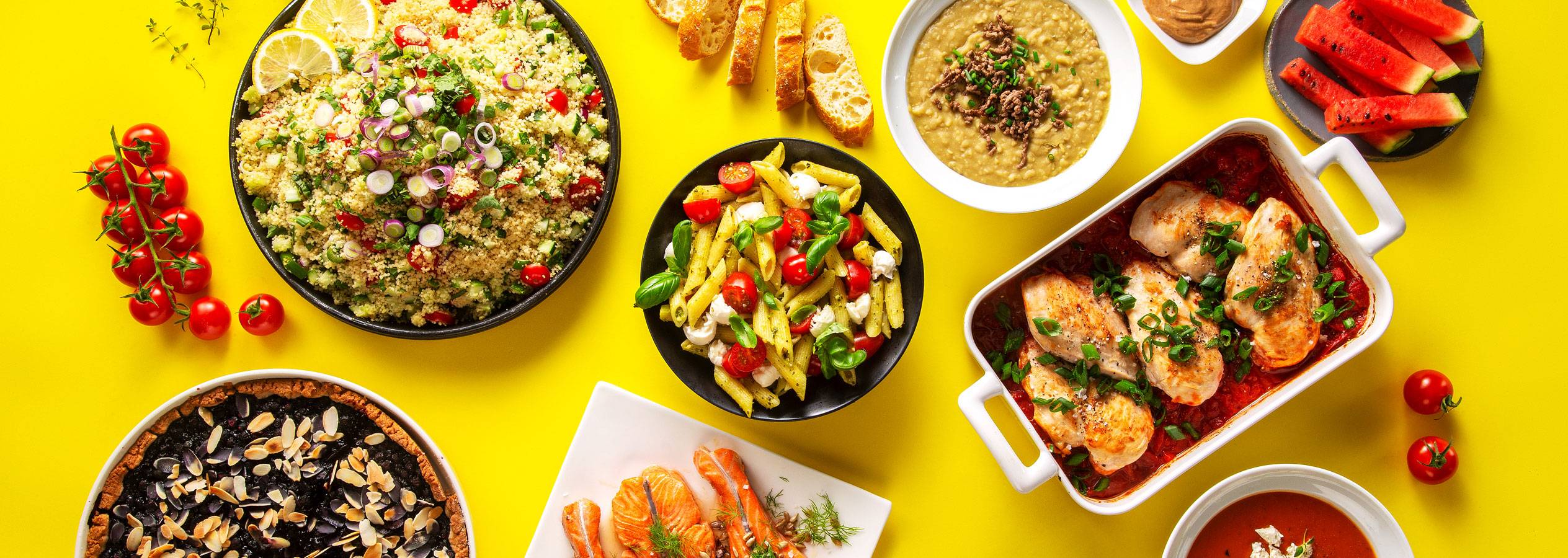 Värikäs valikoima herkullisia ruokia keltaisella taustalla: couscous-salaatti, pasta, kana, lohi ja muita herkkuja.