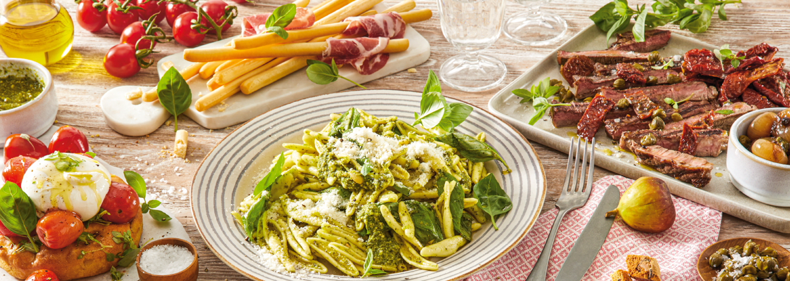 Italialainen ateria, jossa on pesto-pastaa, pihviä, burrataa ja leipätikkuja.