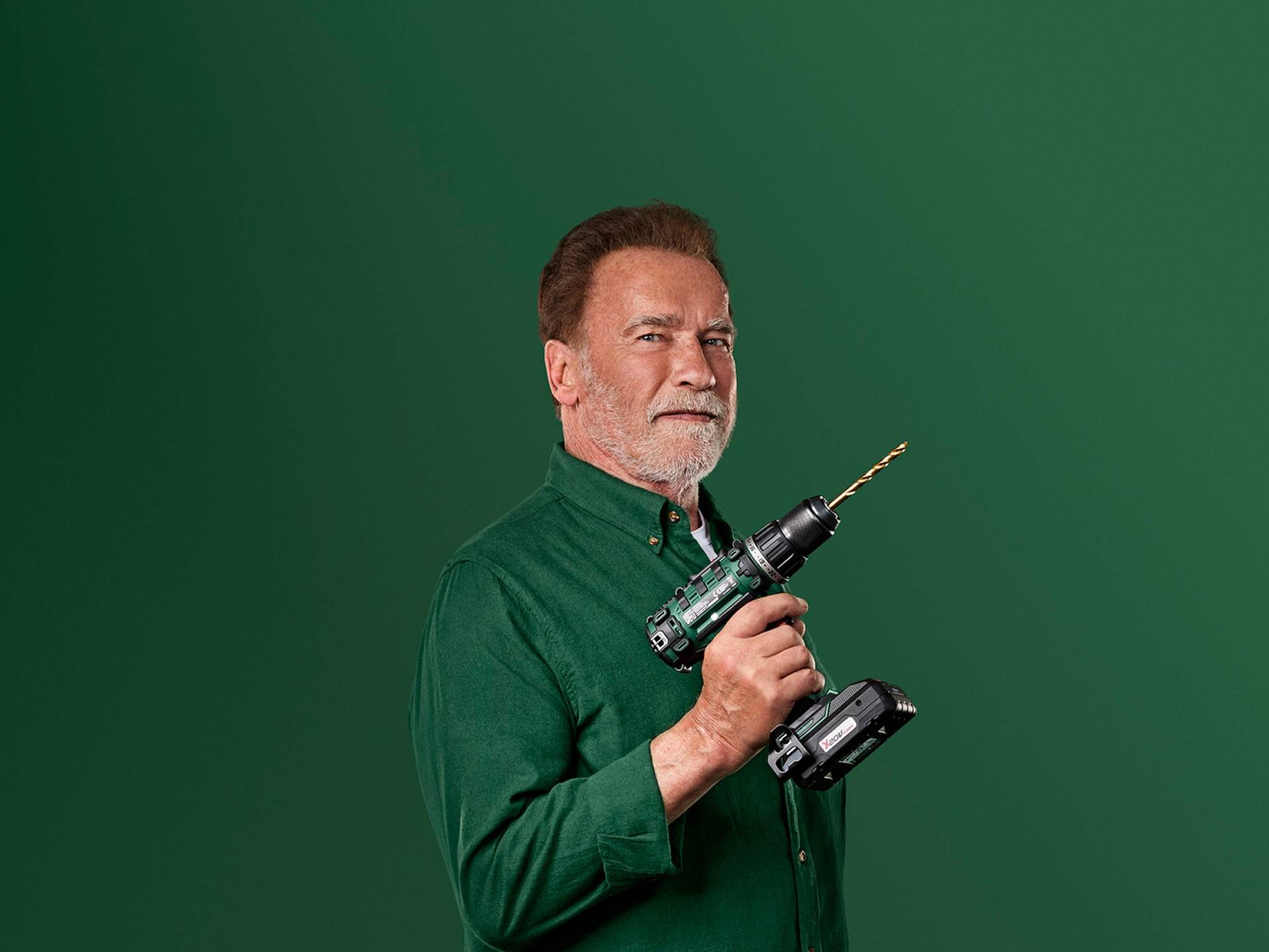 Arnold Schwarzenegger pitää vihreää sähköporaa vihreällä taustalla.