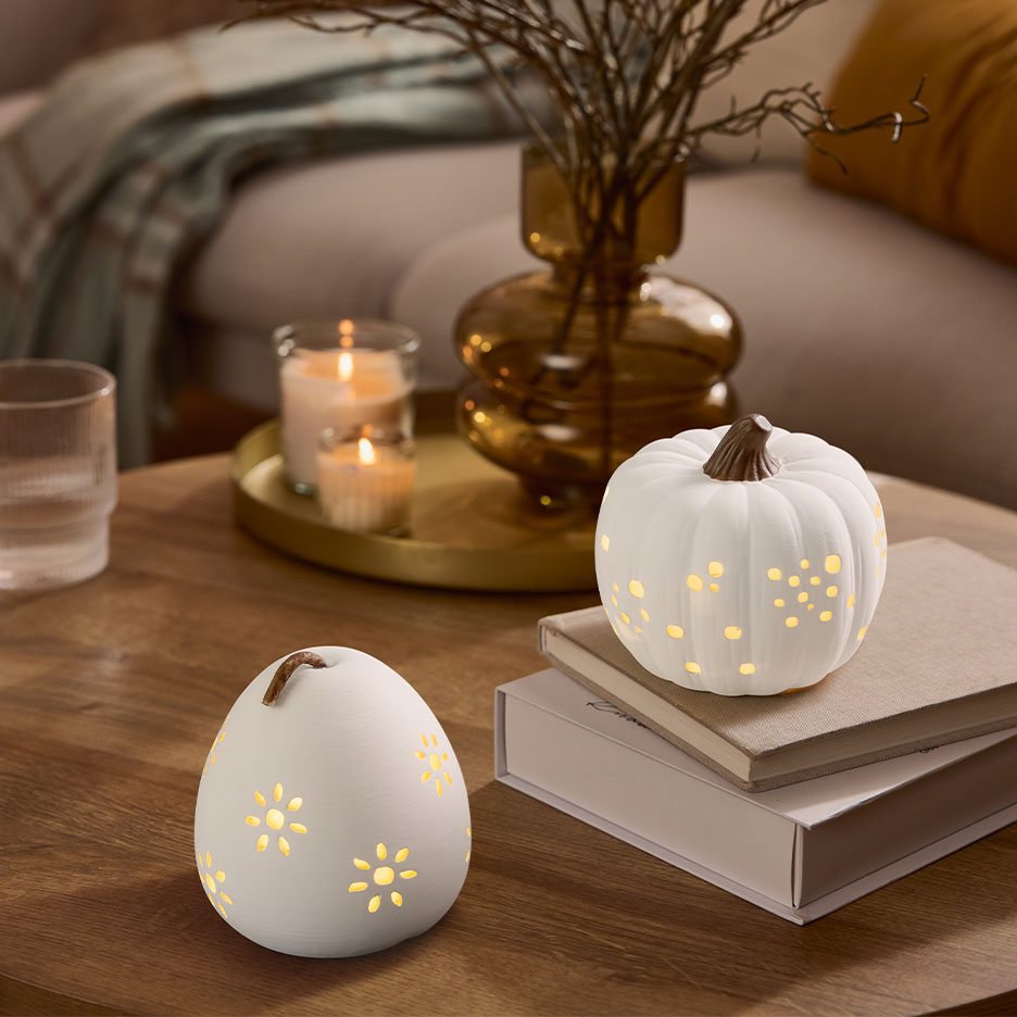 Kaksi valkoista valaistua koristekurpitsaa puupöydällä kynttilöiden ja maljakon kanssa.