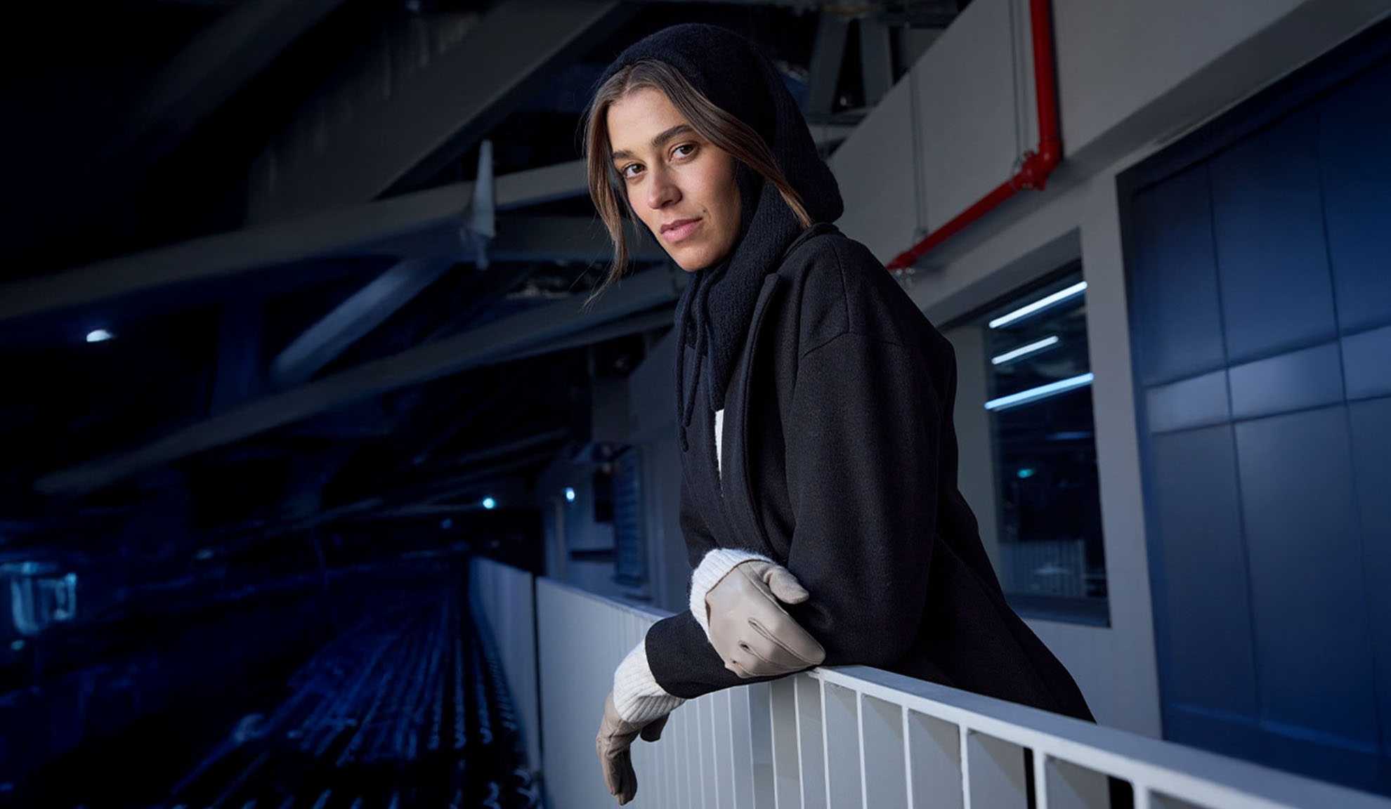 Nainen mustassa takissa ja hupullisessa huivissa, nojaten kaiteeseen modernissa ympäristössä.