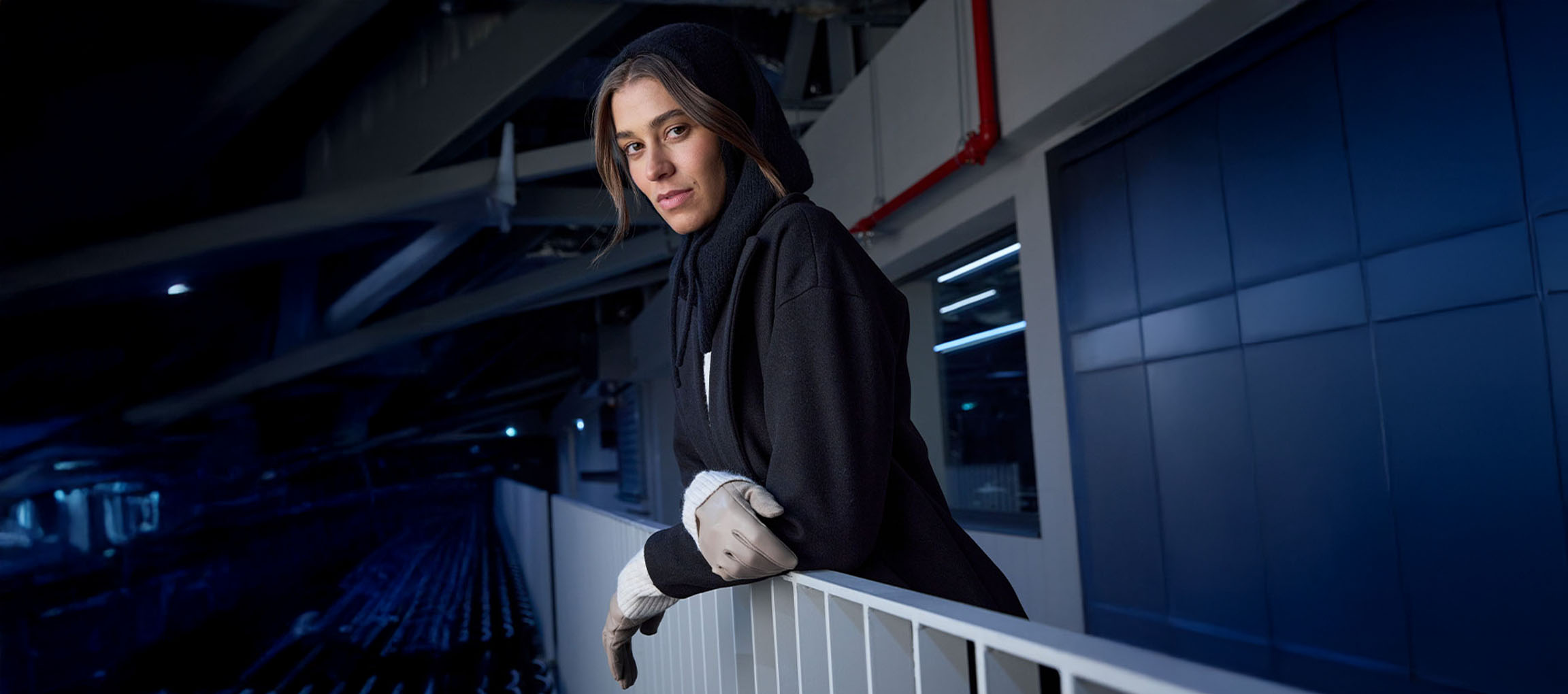 Nainen mustassa takissa ja hansikkaissa nojaa kaiteeseen kaupunkiympäristössä.