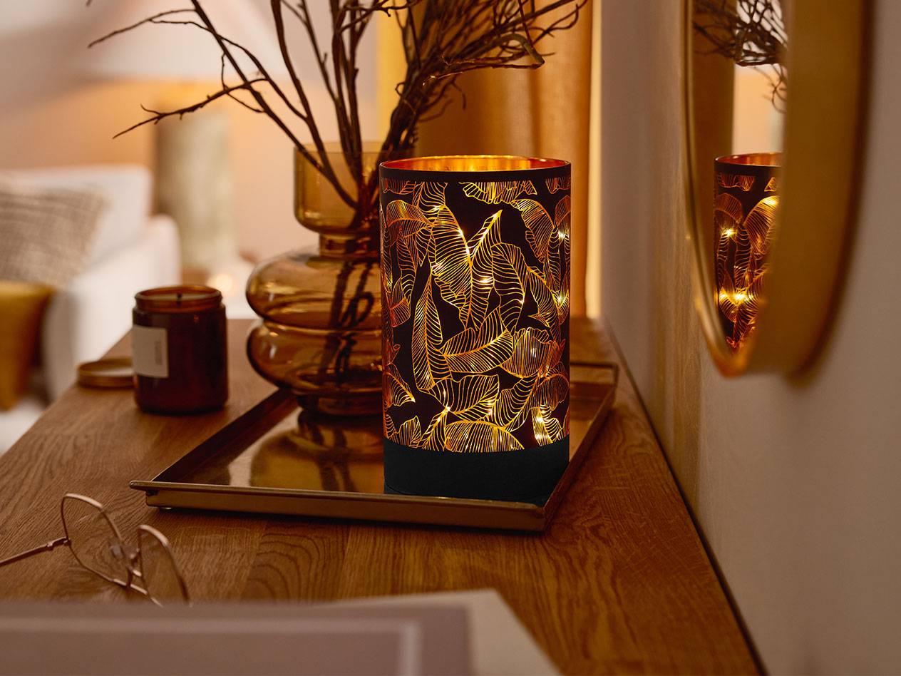 Koristeellinen LED-lyhty lehtikuviolla, maljakko oksineen ja kynttilä puupöydällä.