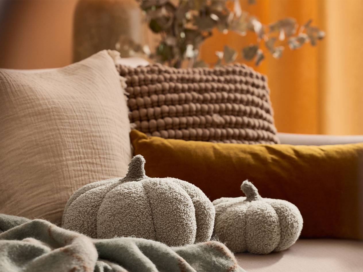 Kaksi pehmeää koristekurpitsaa ja tyynyjä maanläheisissä sävyissä sohvalla.