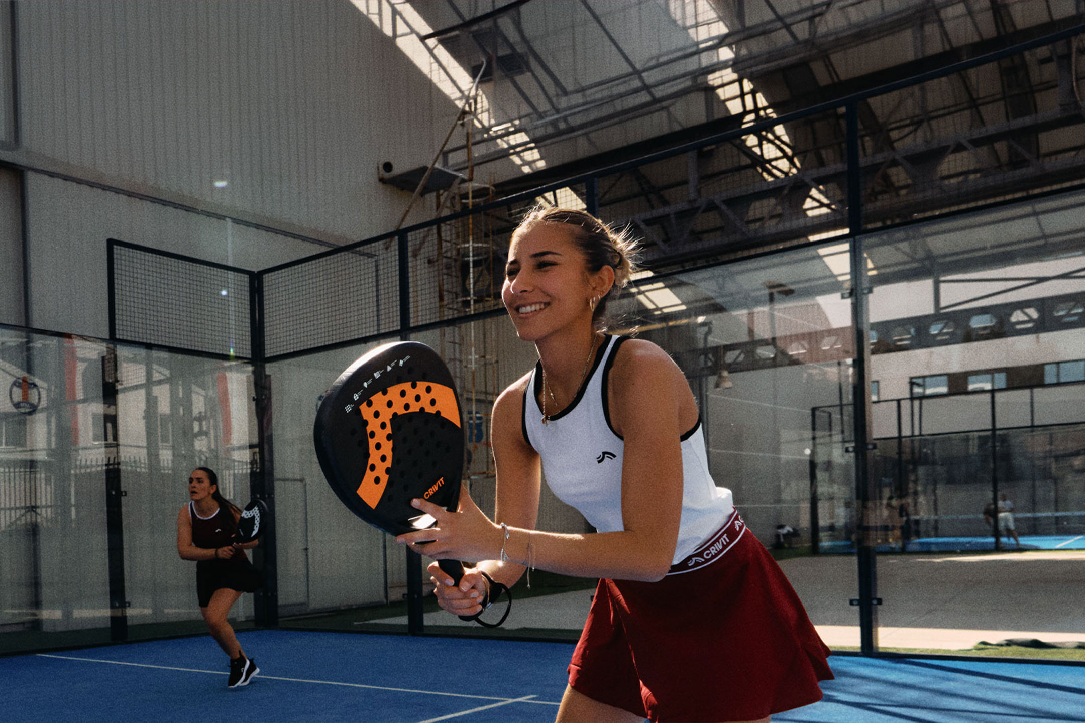 Tekoälyllä luotu: Kaksi naista pelaa padel-tennistä, toinen hymyilee ja pitää mailaa, sinisellä kentällä.