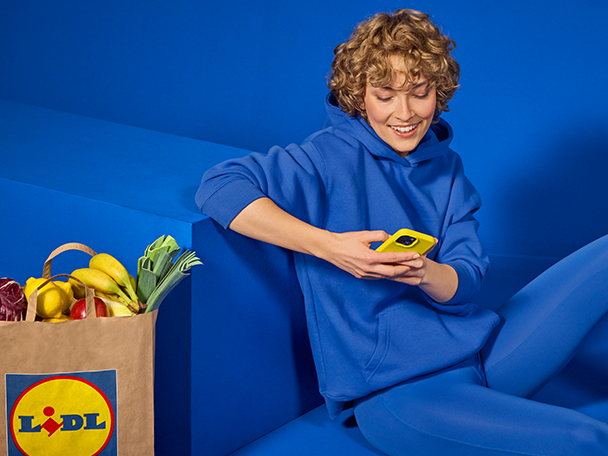 Nainen sinisessä hupparissa selaa puhelinta Lidl-ostoskassin vieressä.