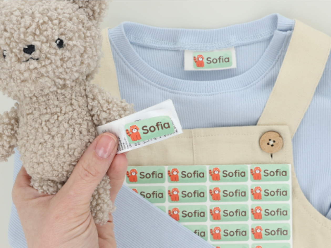 Nimiä tarrat, joissa on punainen panda ja teksti 'Sofia', kiinnitettynä nallekarhuun ja vauvanvaatteisiin.