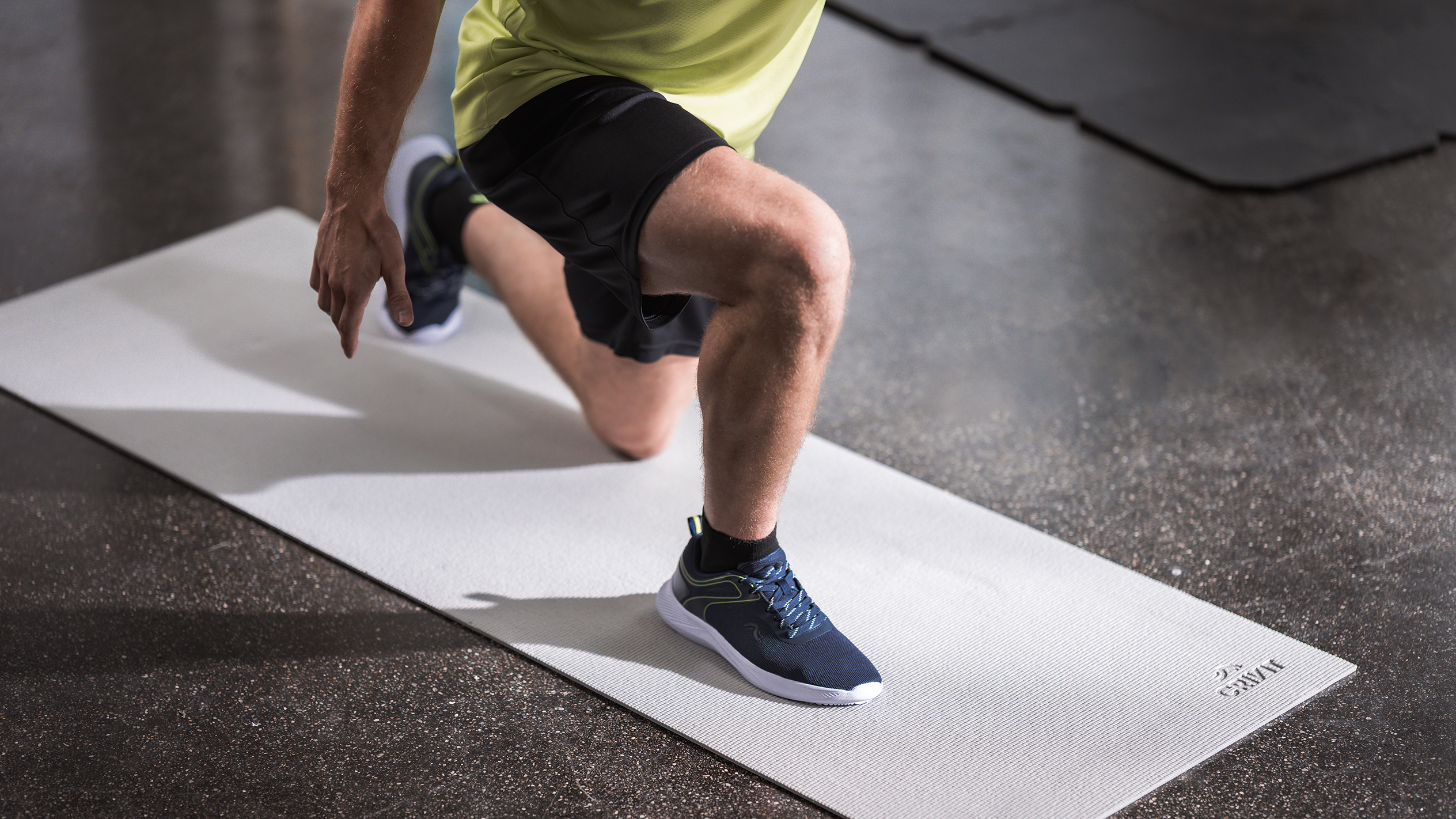 Mies urheiluvaatteissa tekee askelkyykkyjä vaaleanharmaalla jumppamatolla.