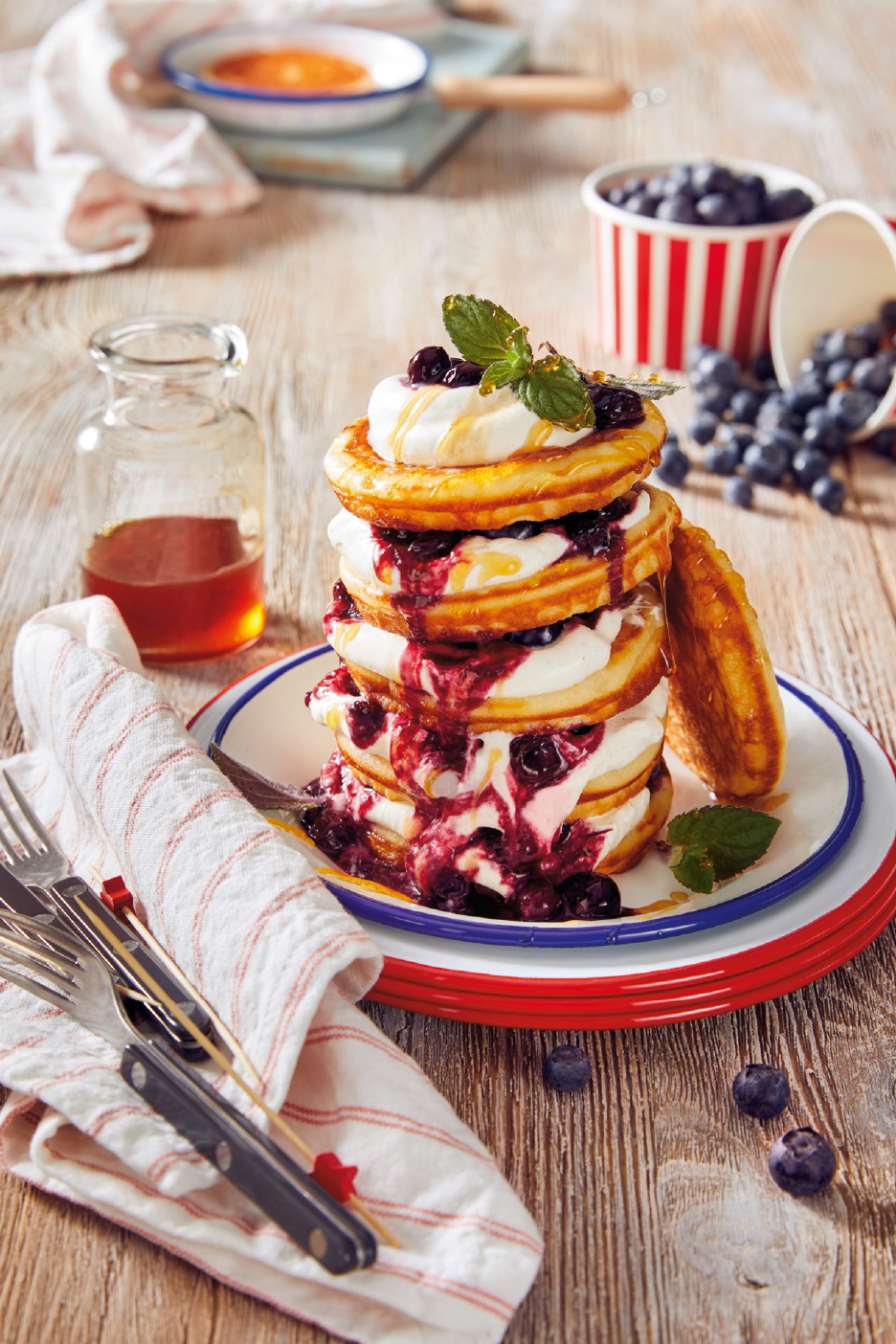 Pannukakkutorni, jossa on kermavaahtoa, mustikoita ja siirappia puupöydällä.