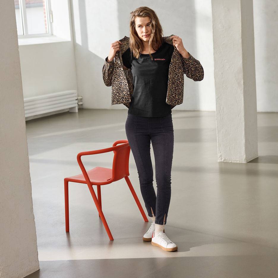 Nainen mustassa t-paidassa, jossa teksti, farkuissa ja leopardikuvioisessa takissa, seisoo punaisen tuolin vieressä.