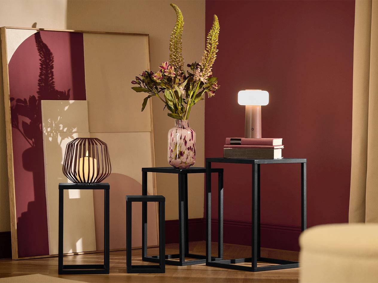 Kolme mustaa sivupöytää kynttilällä, kukkamaljakolla ja lampulla.
