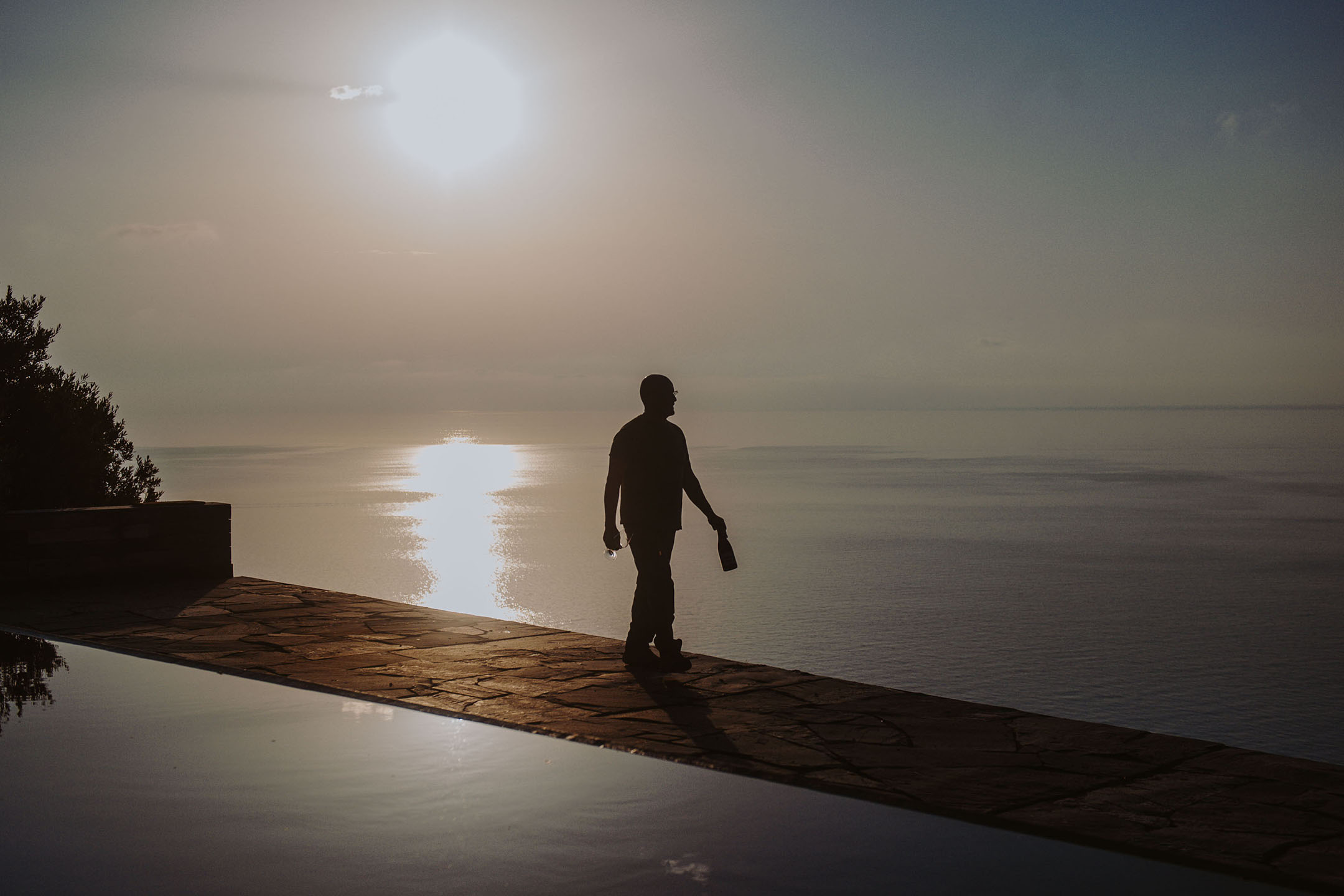 Henkilön siluetti pullon ja lasin kanssa äärettömän uima-altaan äärellä auringonlaskussa