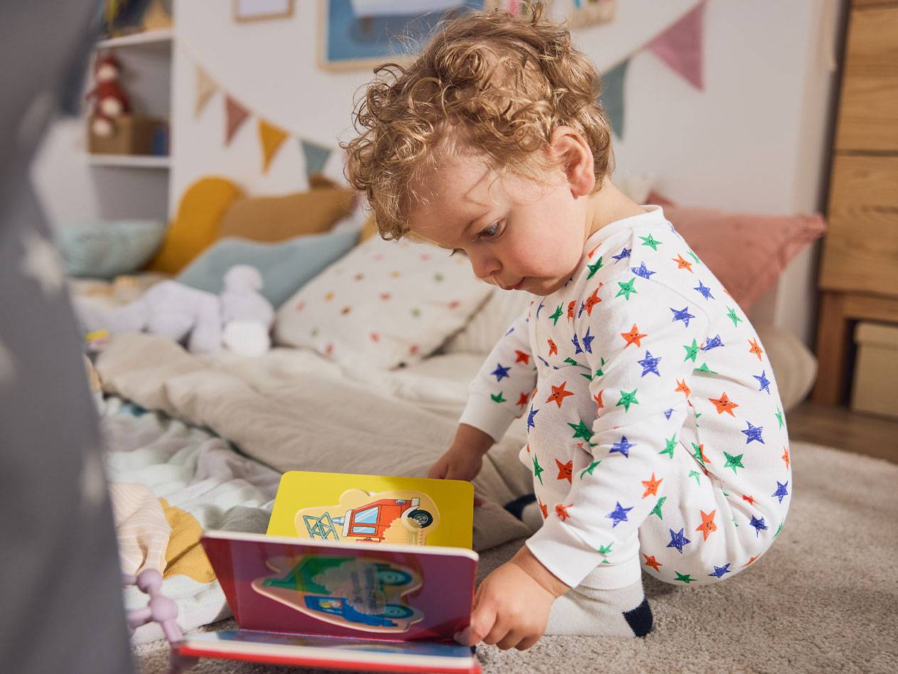 Pieni lapsi tähtipyjamassa, istuu matolla ja lukee kirjaa.