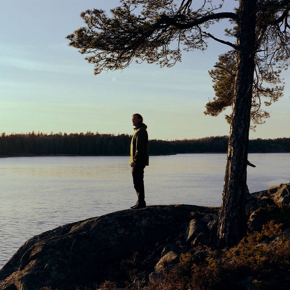 Mies vaellustakissa järven rannalla, puu etualalla.