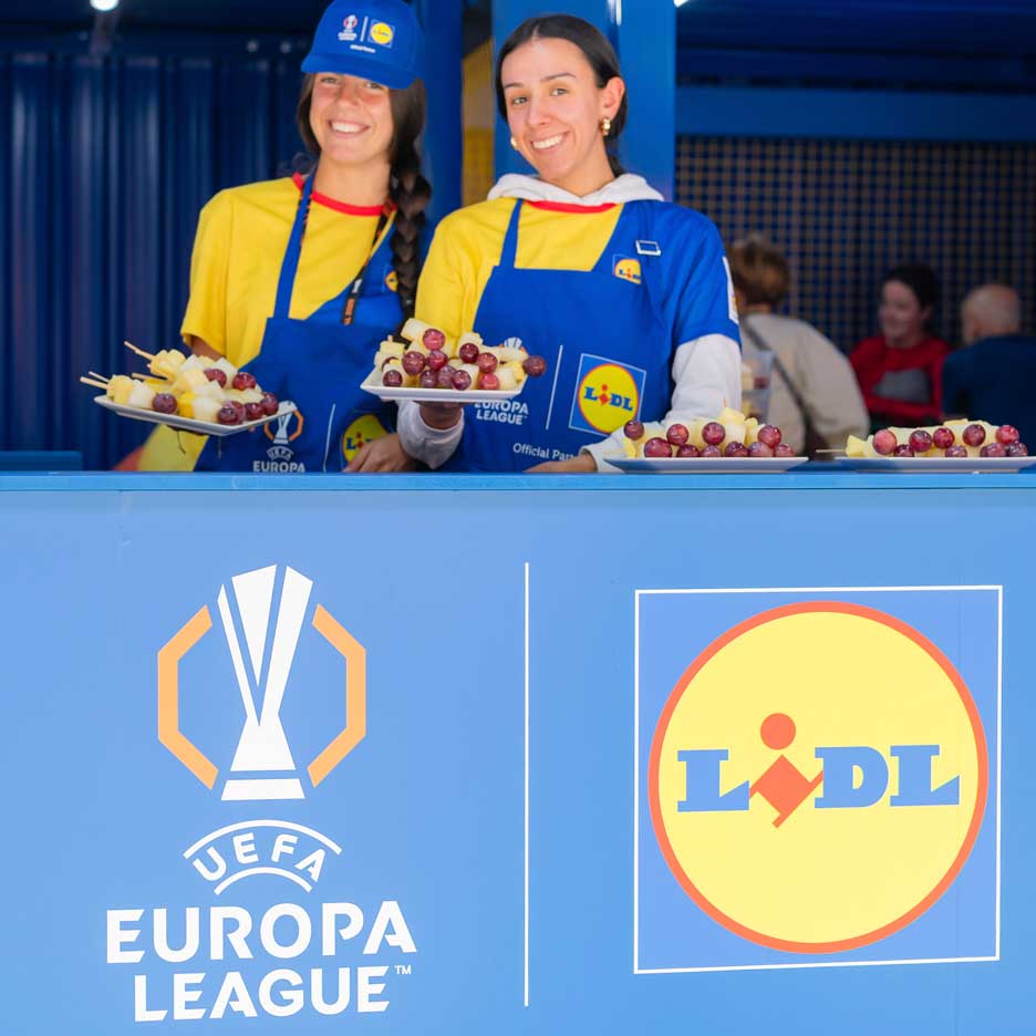 Kaksi hymyilevää naista Lidl-esiliinoissa tarjoaa hedelmiä vartaissa UEFA Europa League -tapahtumassa.