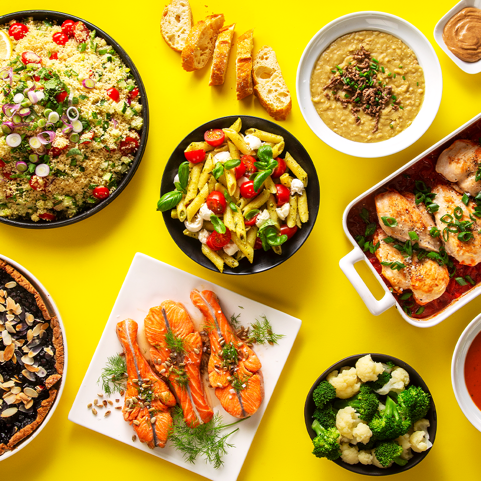 Kattava valikoima ruokia, kuten couscous-salaattia, lohta, kanaa ja pastaa kirkkaankeltaisella taustalla.