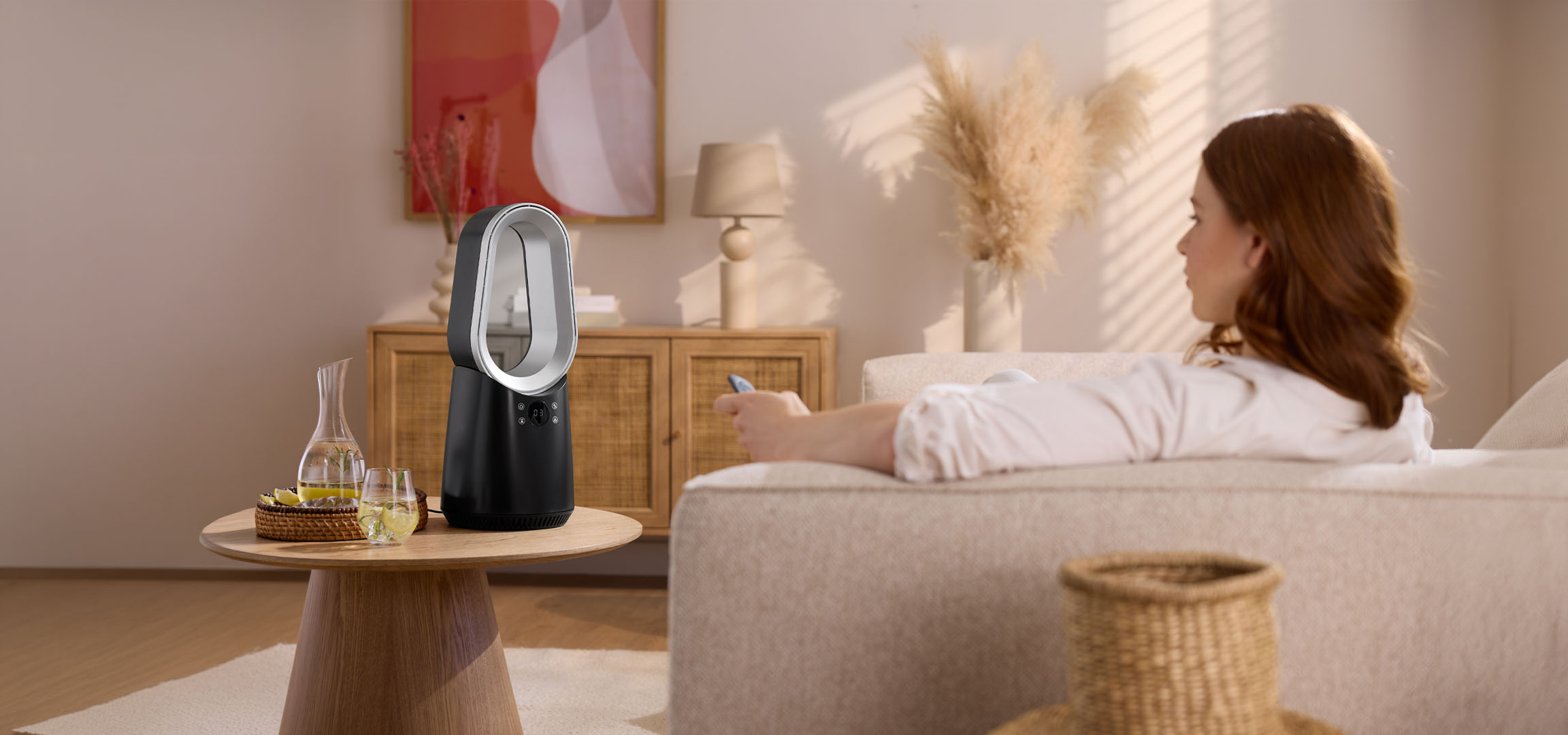 Nainen sohvalla kaukosäätimen kanssa, moderni tuuletin ja juomia pöydällä.