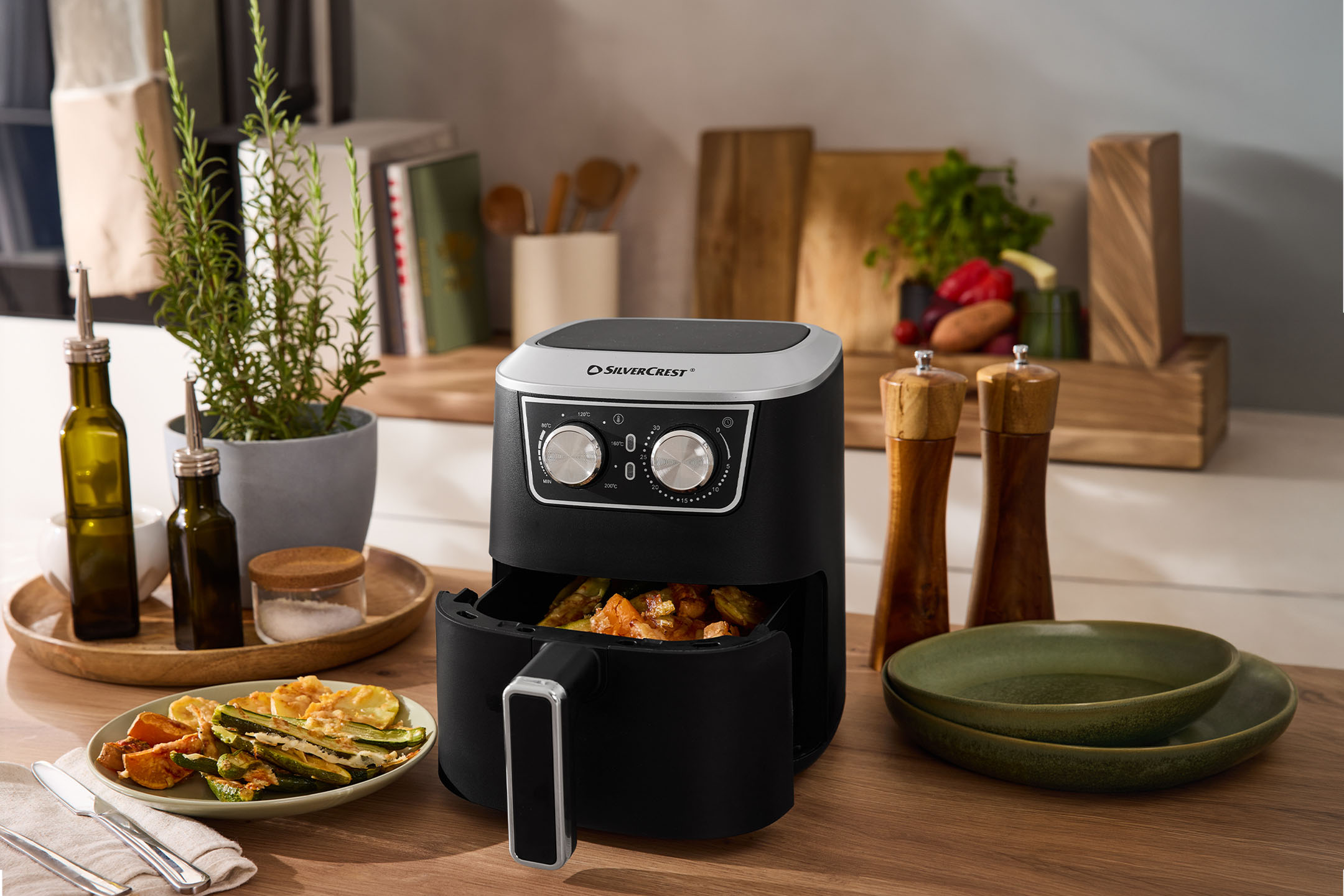 Musta ilmakeitin vihannesten, mausteiden ja astioiden kera puisella työtasolla.