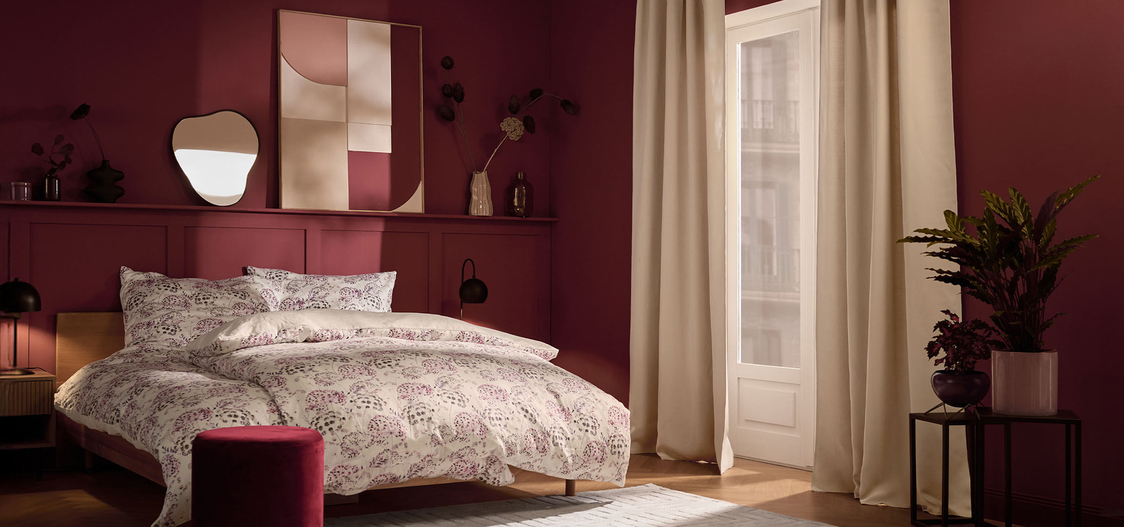 Makuuhuone viininpunaisilla seinillä, sänky valkoisilla kukkakuvioisilla vuodevaatteilla ja kasveilla.