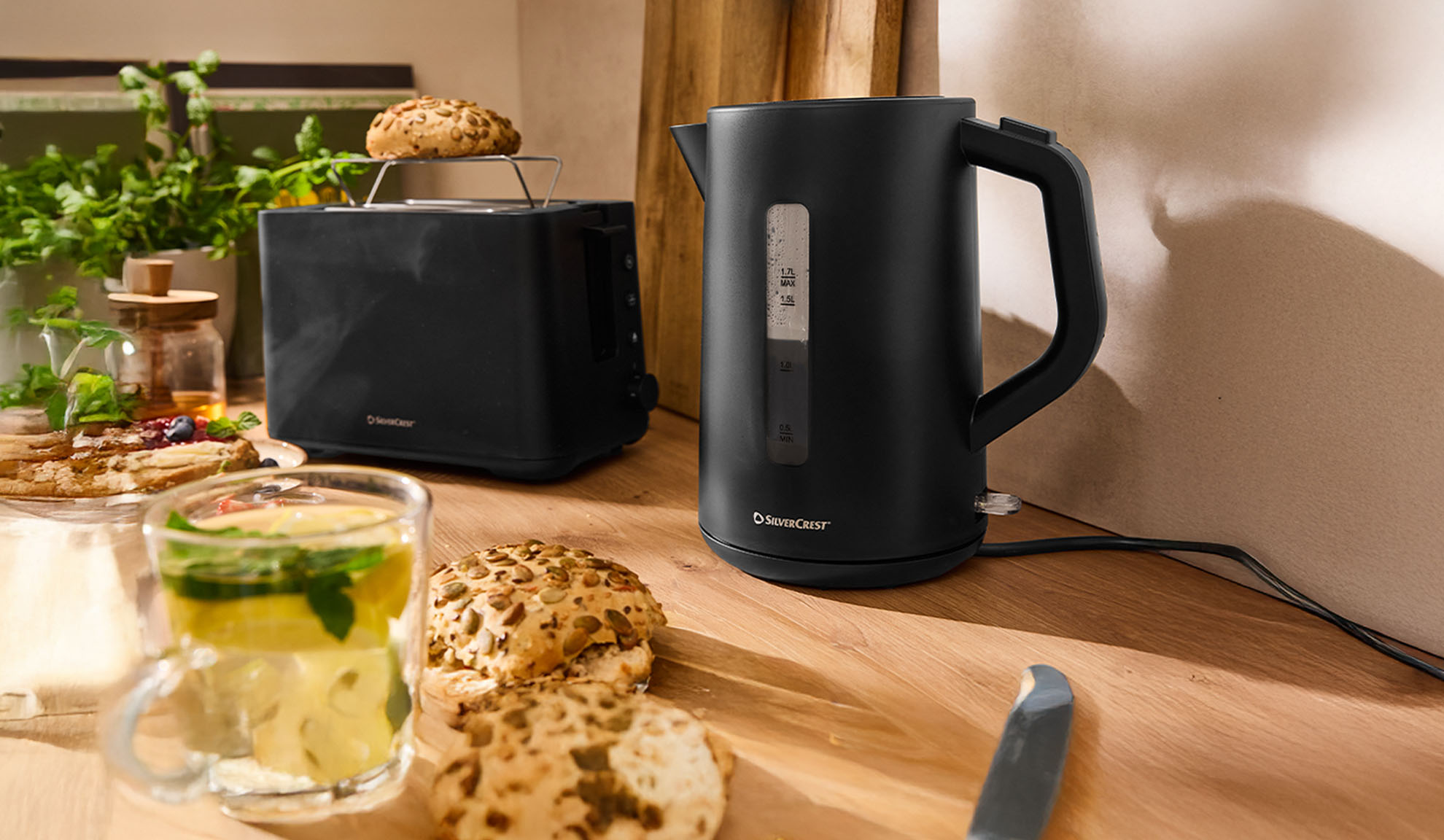 Musta SilverCrest vedenkeitin ja leivänpaahdin puupöydällä aamiaisen kera.