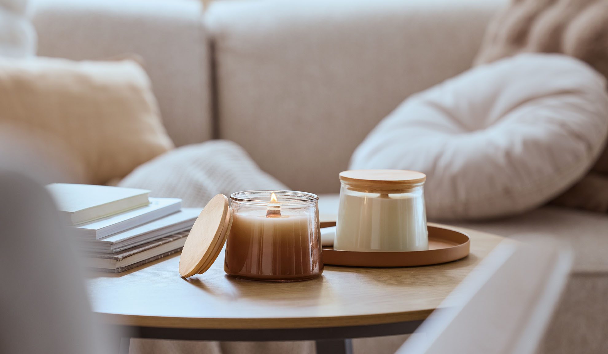 Kaksi tuoksukynttilää puupöydällä, toinen palaa, taustalla viihtyisä sohva.