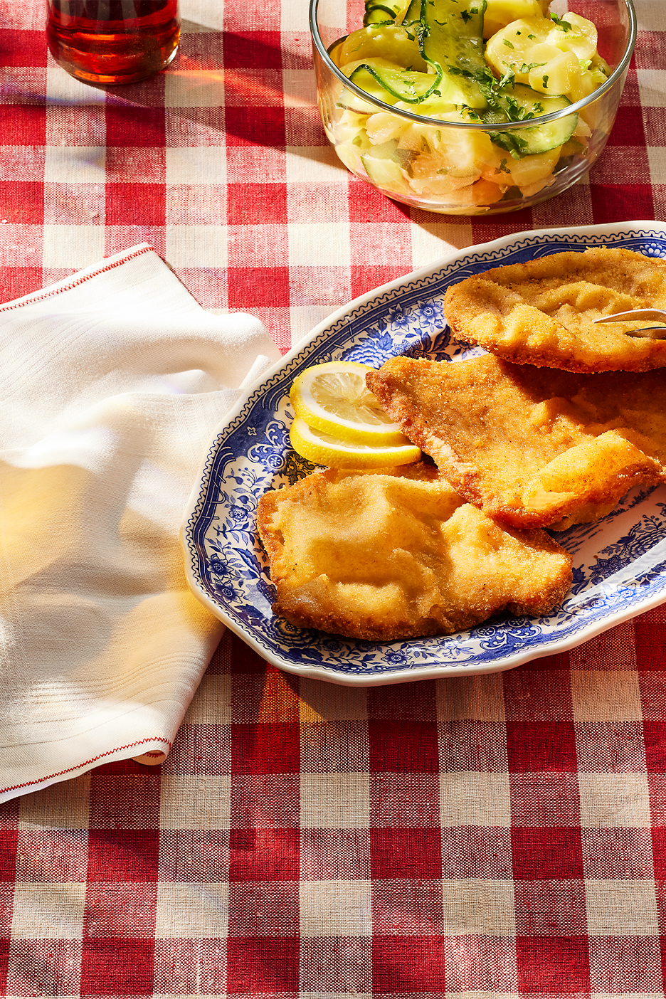 Paneroituja leikkeitä sitruunaviipaleilla, perunasalaattia ja juoma ruudullisella pöytäliinalla.