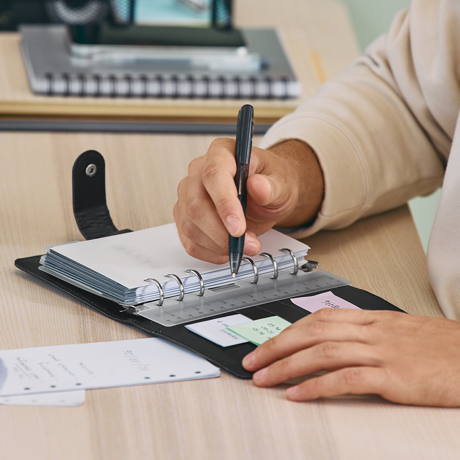 Lähikuva henkilöstä kirjoittamassa mustaan kalenteriin kynällä.