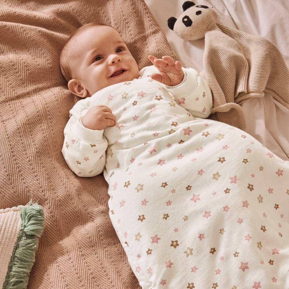 Hymyilevä vauva valkoisessa makuupussissa, jossa vaaleanpunaisia ja ruskeita tähtiä, peiton päällä.