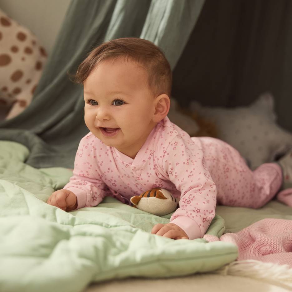 Vauva vaaleanpunaisessa tähtipyjamassa, makaa vihreällä peitolla pehmolelun kanssa.