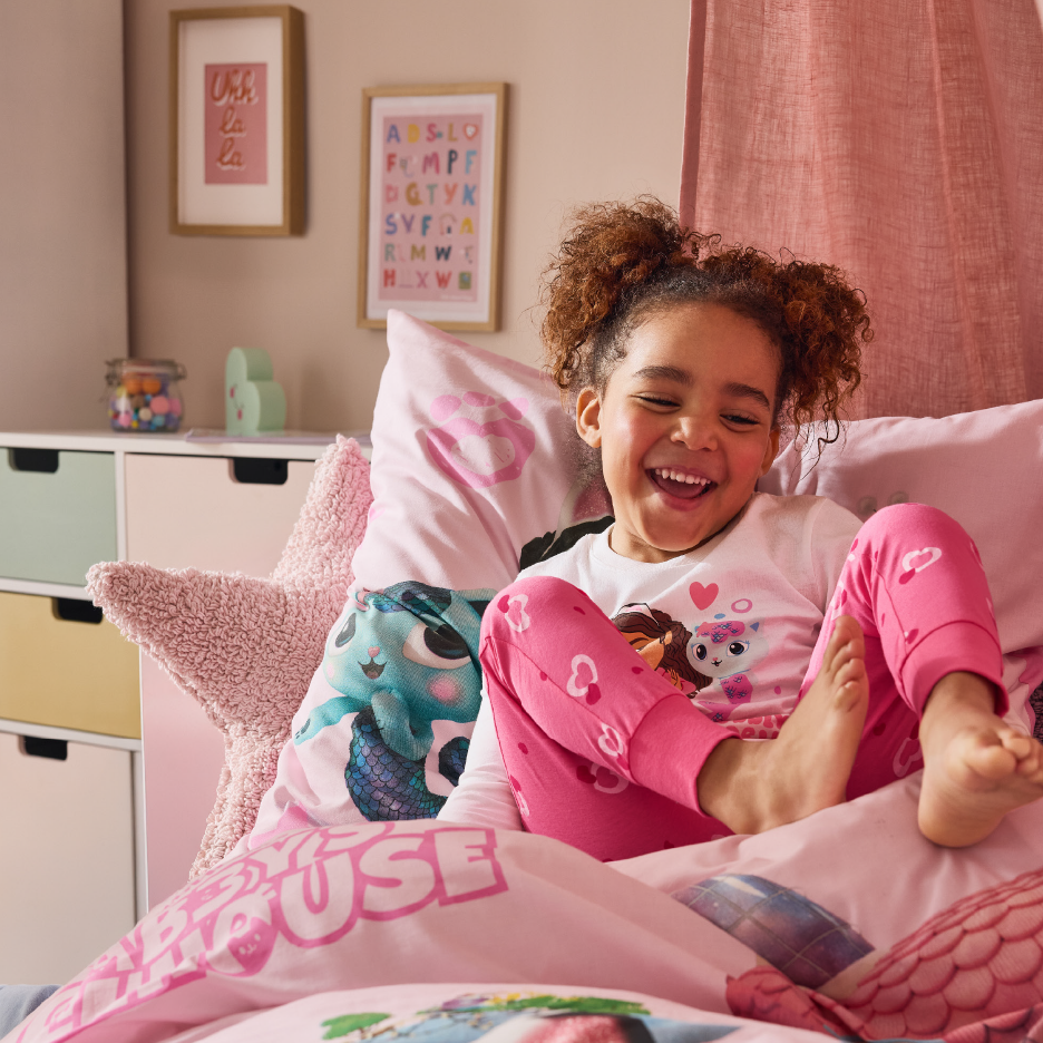 Iloinen tyttö vaaleanpunaisissa Gabby's Dollhouse -pyjamissa sängyssä.