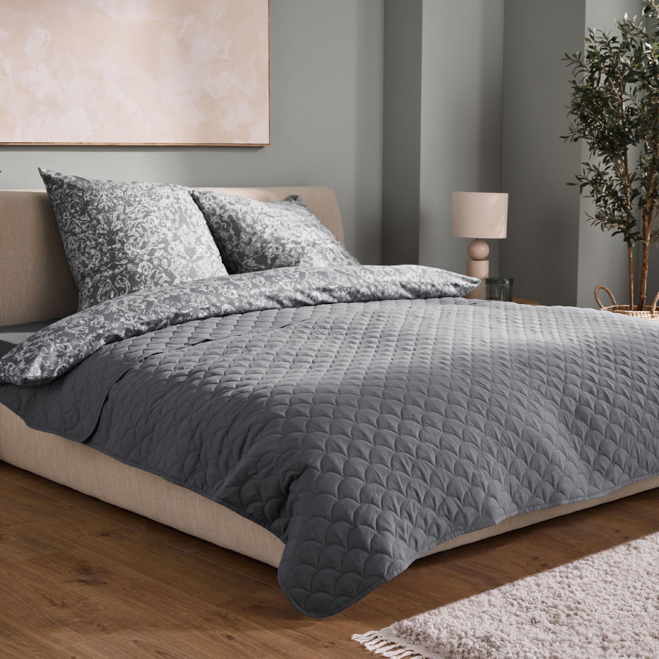 Harmaa tikattu päiväpeitto ja kuviolliset vuodevaatteet makuuhuoneessa.