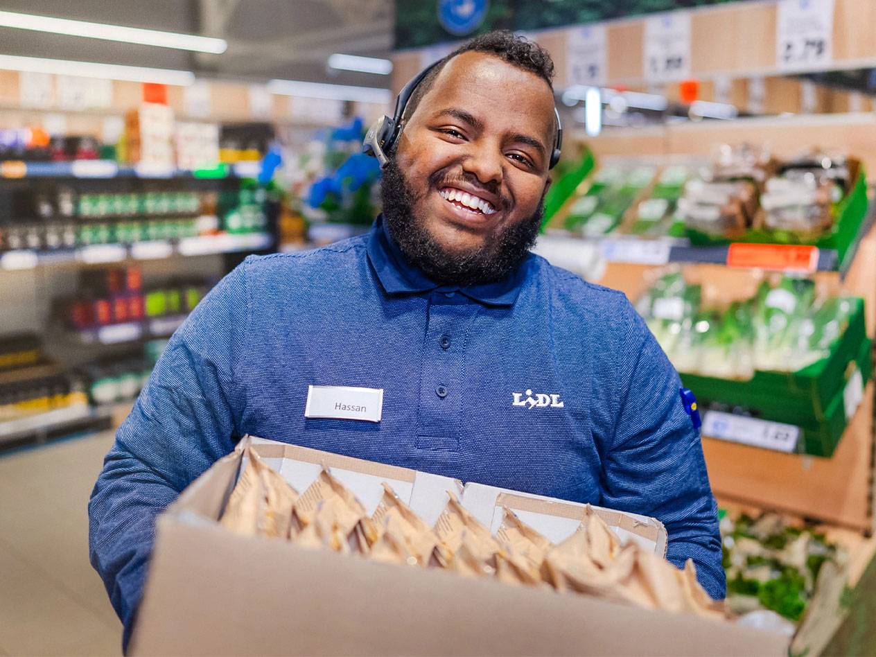 LIDL-työntekijä hymyilee kantaessaan laatikollista tuotteita.