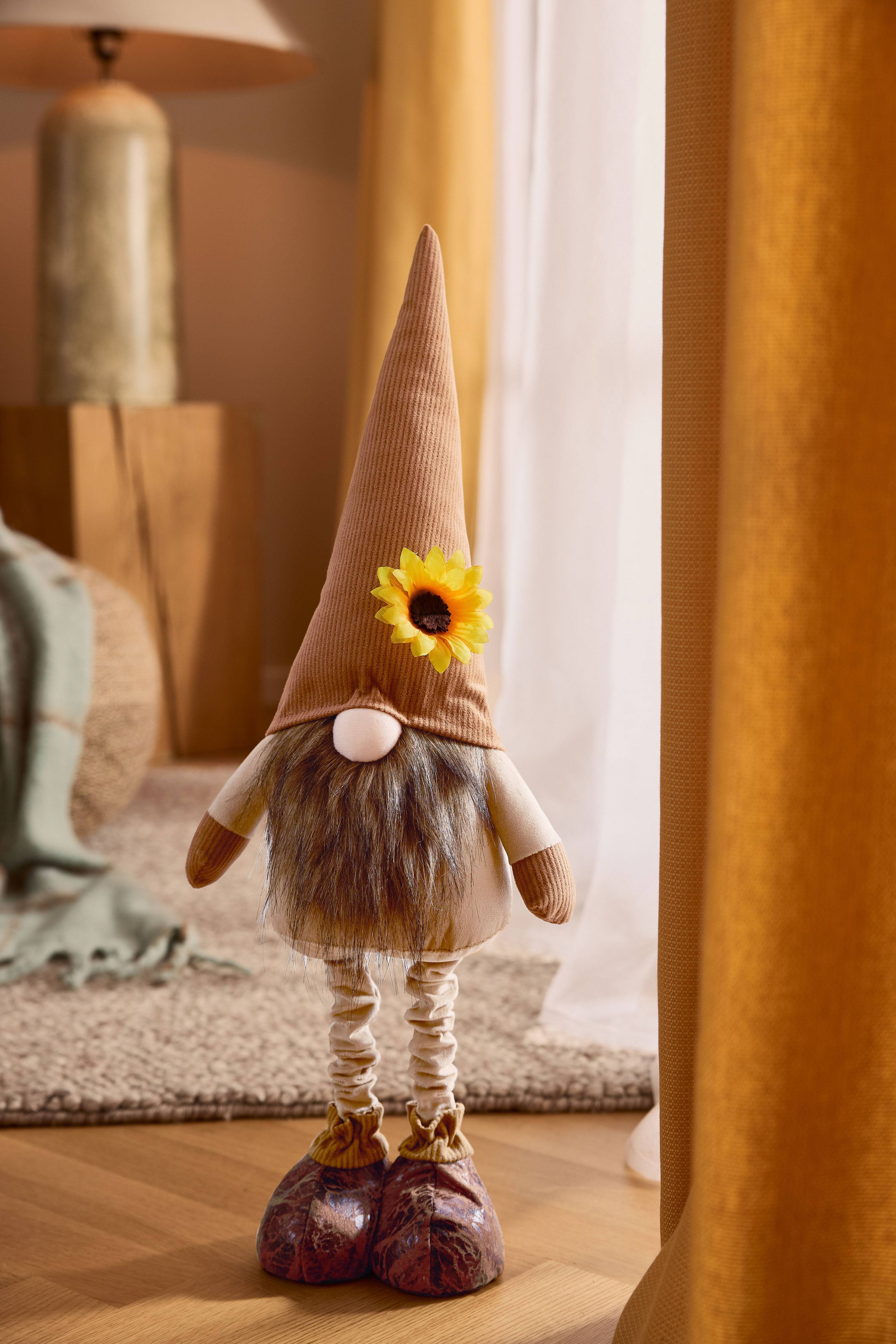 Pehmolelupörröinen tonttu ruskealla suipolla hatulla, parralla ja auringonkukalla, kodikkaassa ympäristössä.