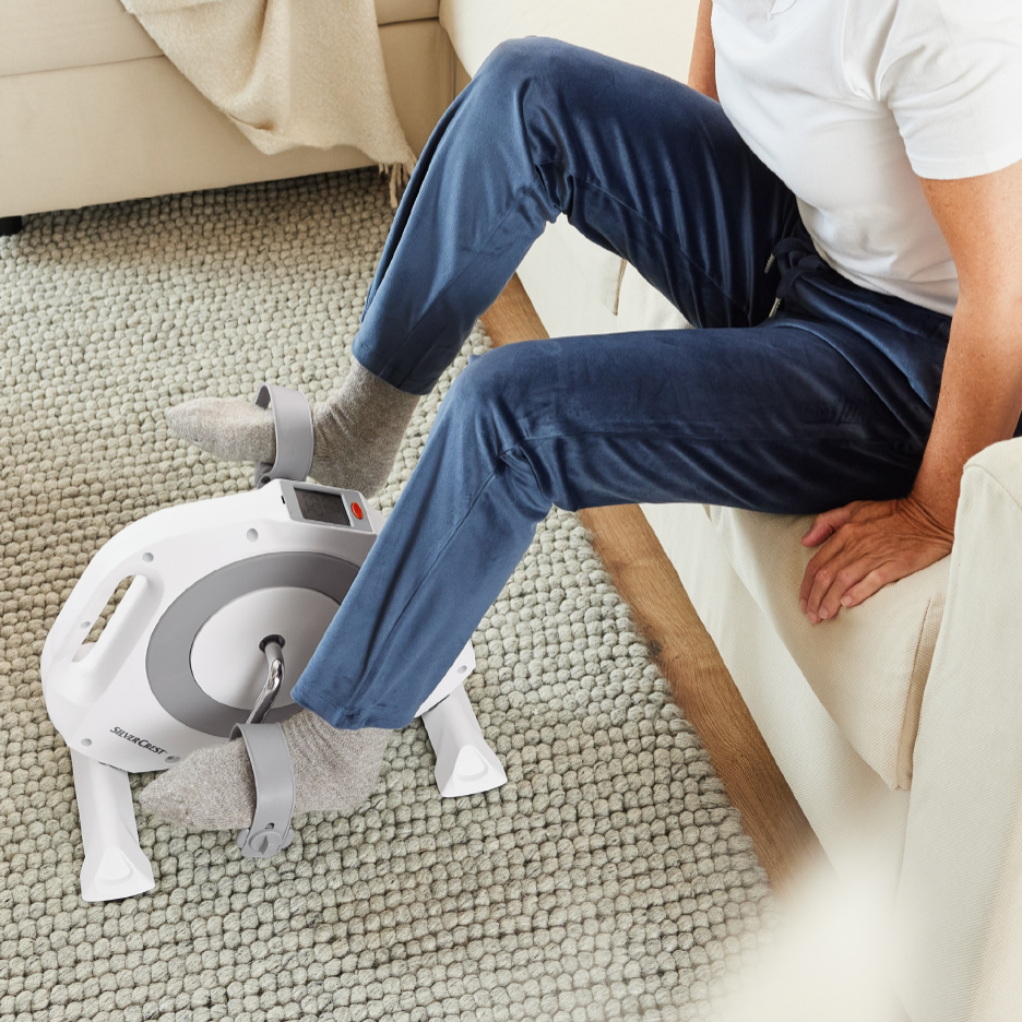 Mies käyttää SilverCrest-polkijalaitetta olohuoneessa sohvalla istuen.