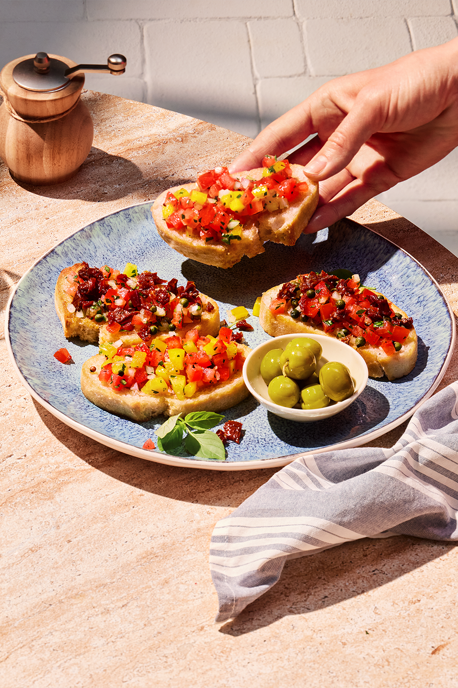 Käsi ottaa bruschettaa tomaatti- ja paprikatäytteellä lautaselta, jossa on myös oliiveja.
