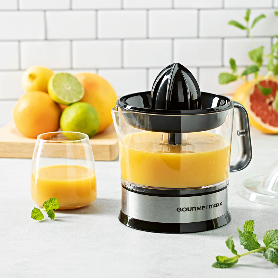 Gourmetmaxx-sitruspuristin täynnä appelsiinimehua, lasi mehua ja sitrushedelmiä taustalla.