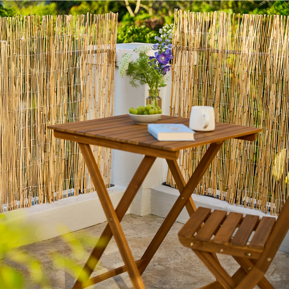 Bambuaidalla rajattu parveke, jossa puinen pöytä ja tuoli, maljakko kukkia ja hedelmiä.