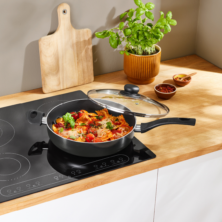 Paistinpannu pastalla induktioliedellä, leikkuulauta ja yrttejä keittiössä.