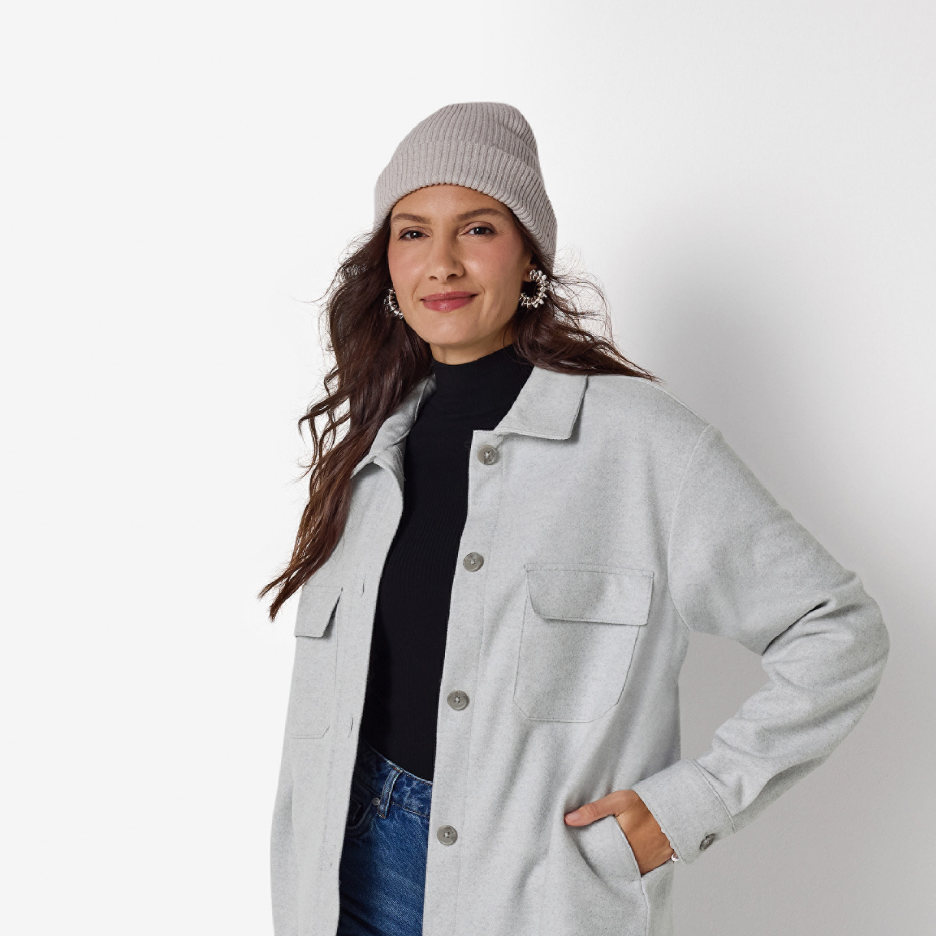 Nainen harmaassa pipossa ja harmaassa shaketissa, musta poolopaita ja farkut.