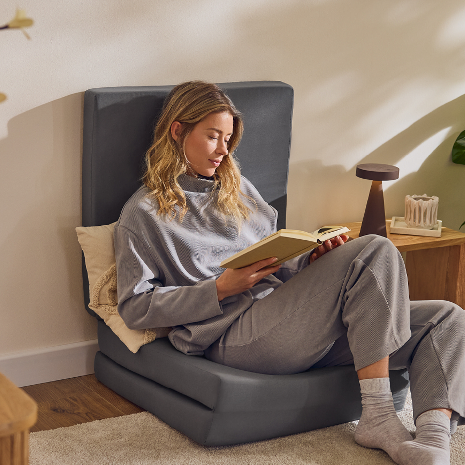Nainen lukee kirjaa harmaalla taitettavalla lattiatuolilla olohuoneessa.