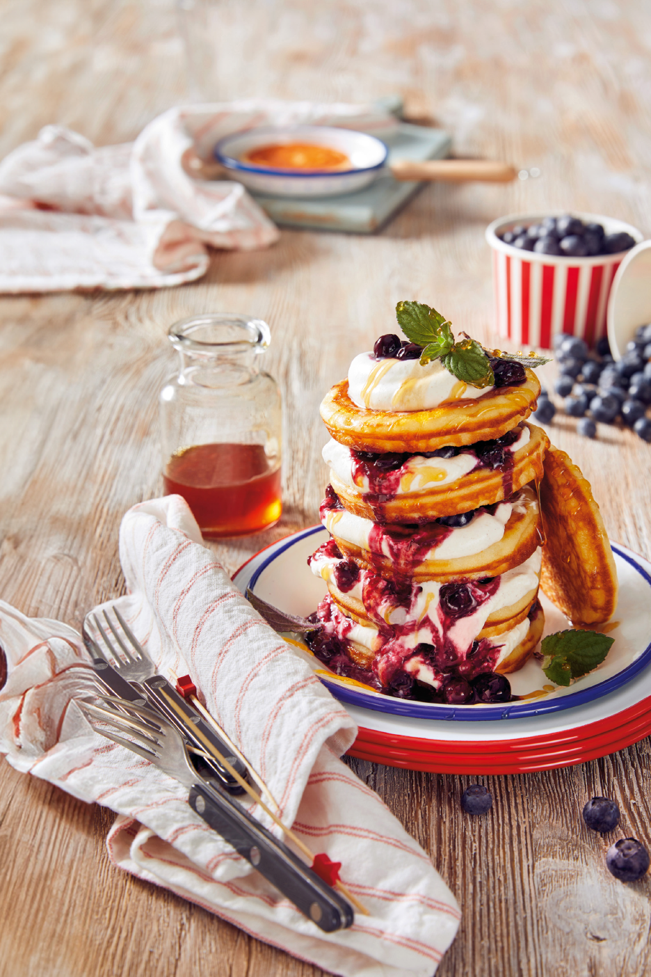 Pannukakkutorni kermavaahdolla, mustikoilla ja siirapilla puupöydällä.