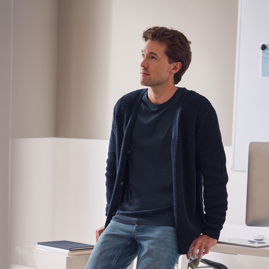 Mies tummansinisessä neuletakissa ja t-paidassa, farkuissa, nojaa pöytään.