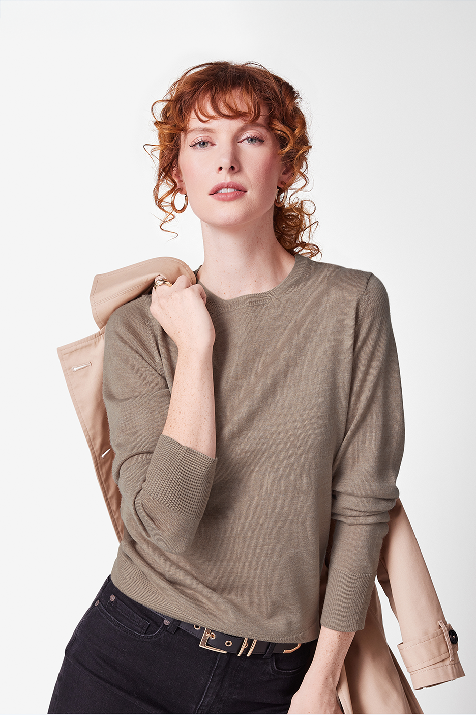 Nainen harmaassa neuleessa, mustissa farkuissa ja beigessä takissa olalla.