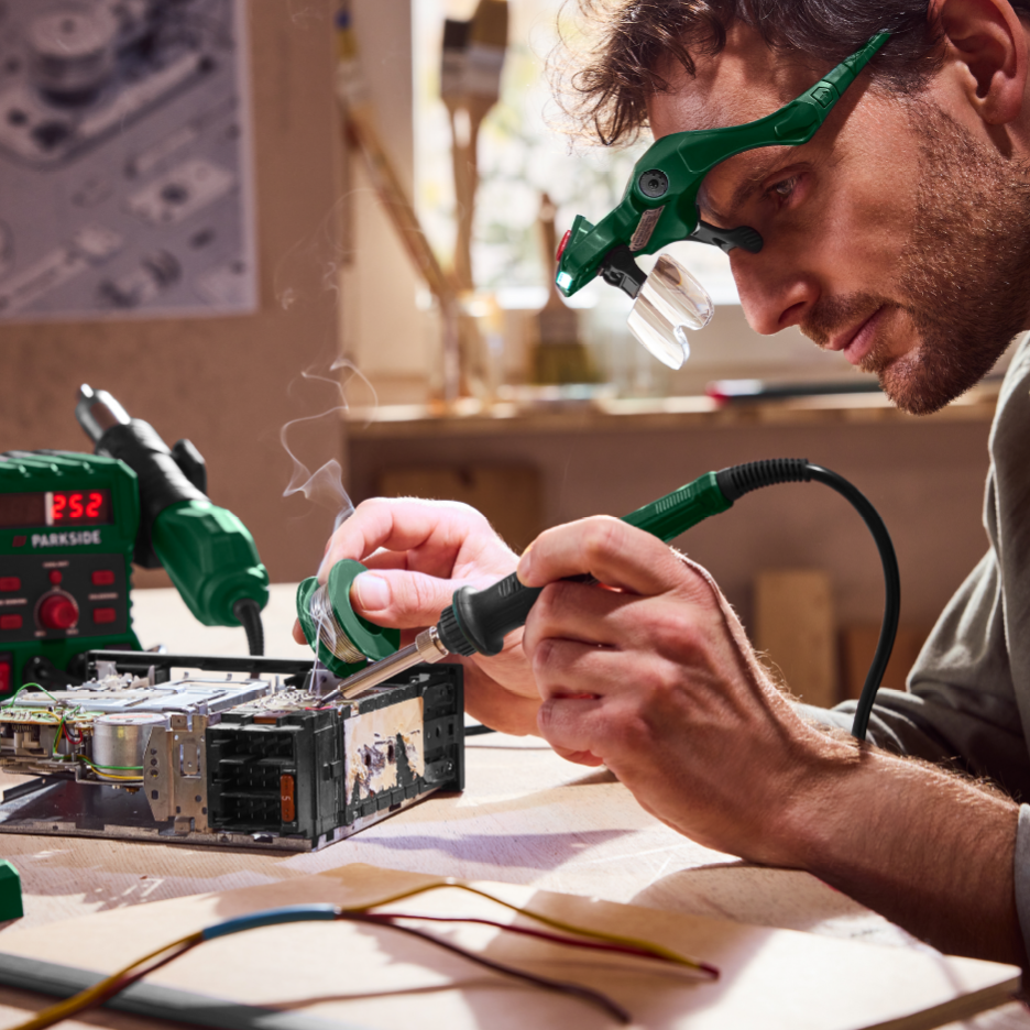 Mies juottaa elektroniikkaa Parkside-juotosasemalla ja suurennuslasilaseilla.