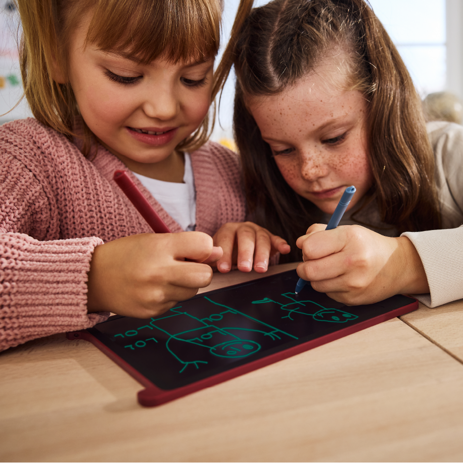 Kaksi tyttöä piirtää yhdessä punaiselle piirustusalustalle, toinen piirtää punaisella ja toinen sinisellä kynällä.