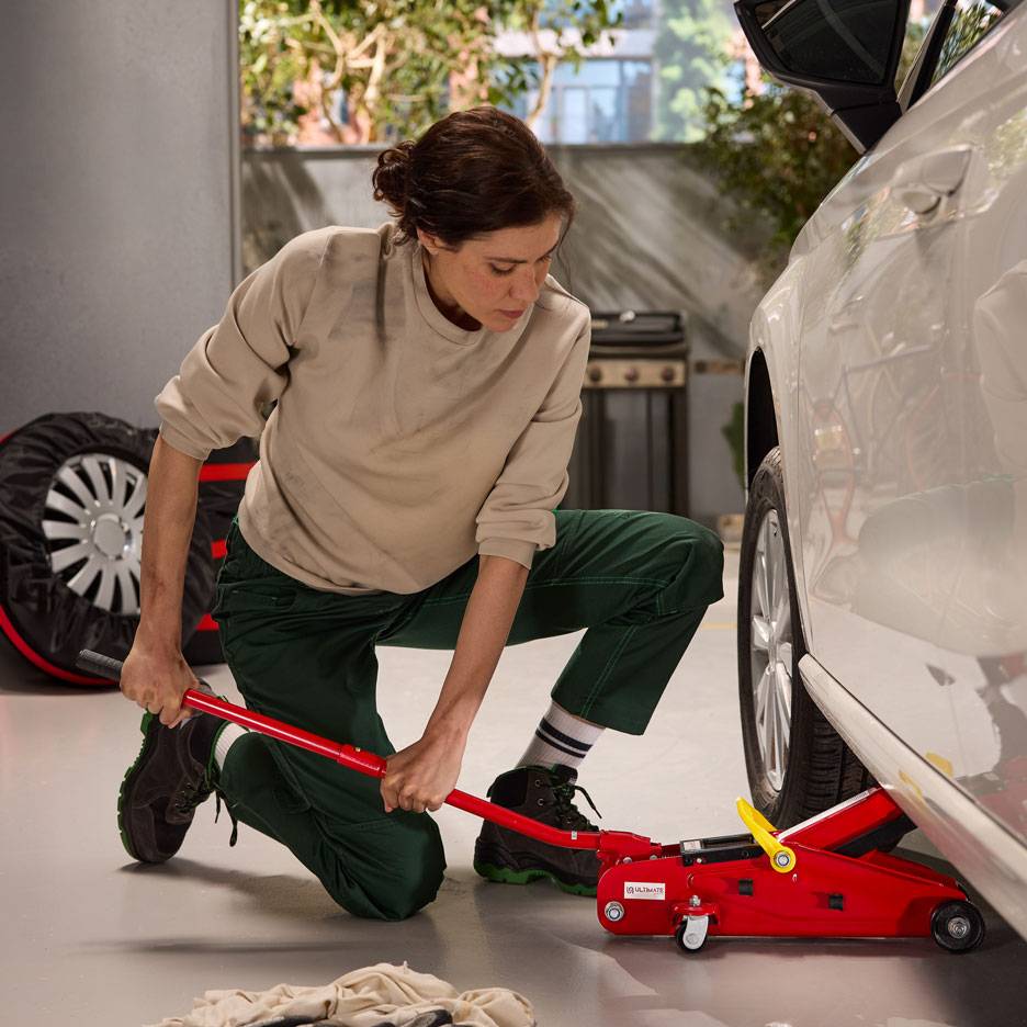 Nainen käyttää punaista hydraulitunkkia nostaakseen valkoista autoa, vararengas lähellä.