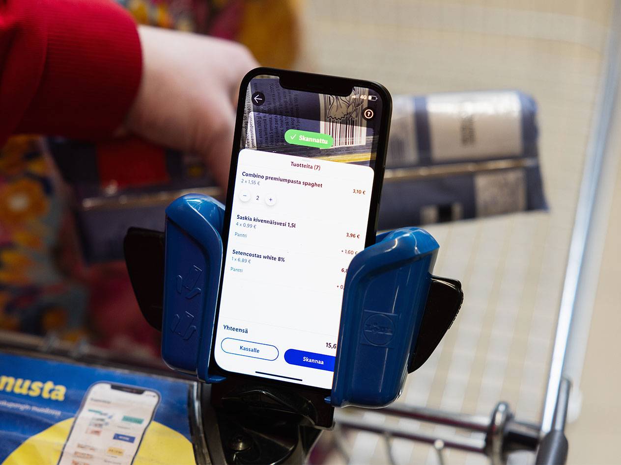 Asiakas skannaa tuotteita ostoskärryssä olevalla puhelimella, näytöllä näkyy pasta, kivennäisvesi ja valkoviini.